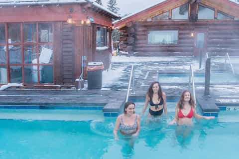 Women in outdoor hot spring pool with rustic log cabin buildings in snowy mountain setting