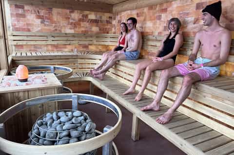 Sauna interior with four people sitting on wooden benches beside stone heater and hot tub