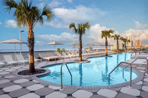 Luxury beachfront pool deck with palm trees, umbrellas, loungers, and ocean city skyline