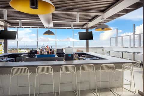 Modern rooftop bar with ocean views, yellow heaters, white bar seating, flat-screen TVs, and horizon view over waterfront