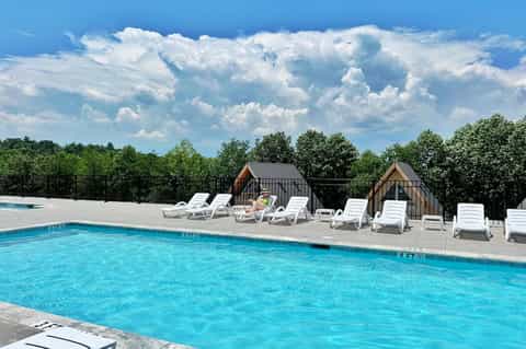 Resort pool with crystal blue water, white lounge chairs, two pool cabanas, and tree-lined background