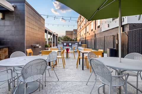 Urban rooftop patio with white tables, yellow chairs, green shade sail, and string lighting