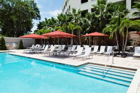 Resort pool with clear blue water, white lounge chairs, red umbrellas, and palm trees surrounding the deck
