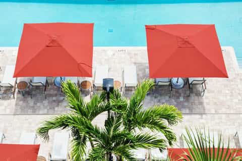 Aerial view of bright turquoise pool with red umbrellas, white lounge chairs, and palm trees