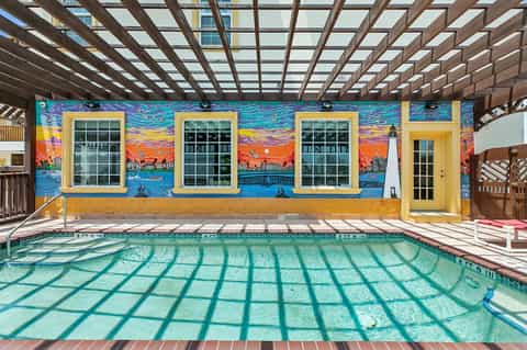 Indoor pool with geometric shadow patterns and vibrant yellow building with painted ocean and sunset mural backdrop