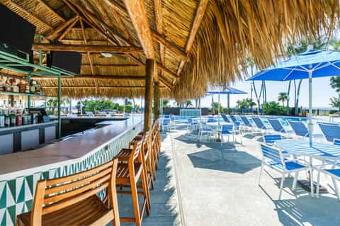 Tiki-style beach bar with thatched roof, wooden chairs, blue umbrellas, and waterfront palm tree setting