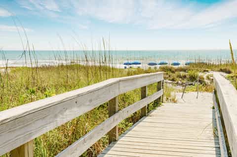 Wooden boardwalk leading to sandy beach with dune grass and blue umbrellas beyond the shoreline