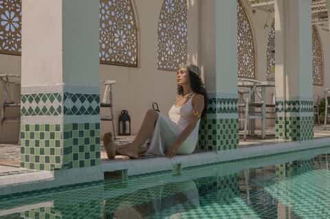Woman in white dress lounging by decorative tiled pool with Moroccan-style architectural details