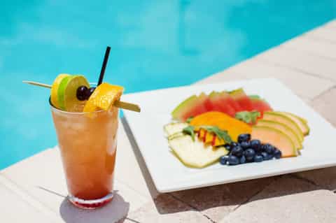Tropical cocktail with fruit garnish beside pool-side fruit plate with berries and melon