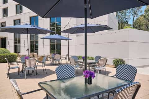Outdoor patio with black umbrellas, striped chairs with purple cushions, potted flowers, and modern hotel building.
