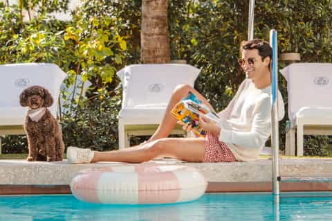 Man relaxing by pool reading magazine with pet dog nearby and lounge chairs under shade