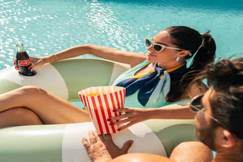 Woman relaxing in pool with colorful floats, sunglasses, holding Coca-Cola and popcorn