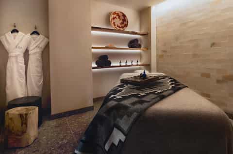 Spa treatment room with massage table, white spa robes, wooden shelves with amenities, and neutral stone tiling