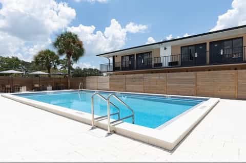 Rectangular swimming pool with white deck, metal railings, palm trees, wooden fence, and two-story building with balconies