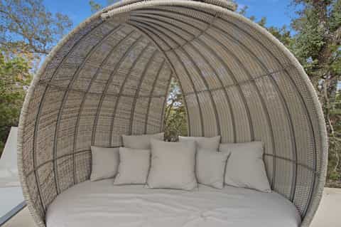 Woven rattan pod-shaped outdoor seating with curved dome, beige cushions, set in landscaped garden
