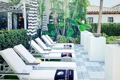 Exclusive resort courtyard with white loungers, striped umbrellas, green privacy hedges, and Spanish colonial architecture.