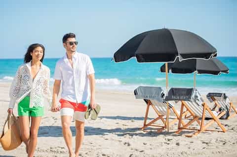 Couple walking on sandy beach with black umbrellas and beach chairs labeled Palm Beach