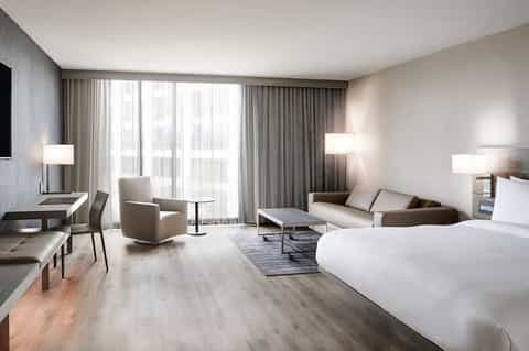 Spacious hotel suite with king bed, seating area, floor-to-ceiling windows with sheer and dark curtains, modern furniture