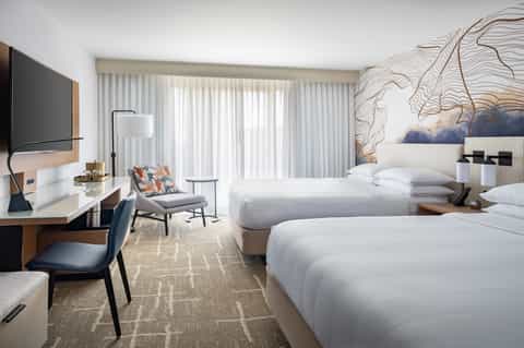 Contemporary hotel room with two queen beds, wall-mounted TV, desk, and floor-to-ceiling windows