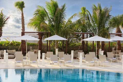 Beachfront resort pool deck with palm trees, white umbrellas, lounge chairs, and ocean horizon backdrop