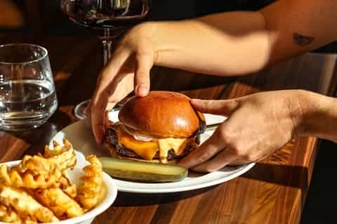 Hands holding gourmet cheeseburger with fries and wine glass on wooden table