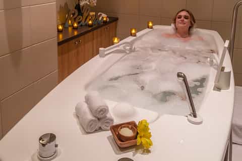 Woman relaxing in spa tub with bubbles, candles, rolled towels, and relaxation signage
