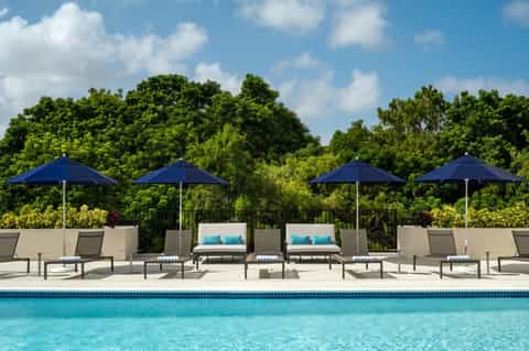 Resort pool deck with lounge chairs, blue umbrellas, daybed seating, and green landscape backdrop