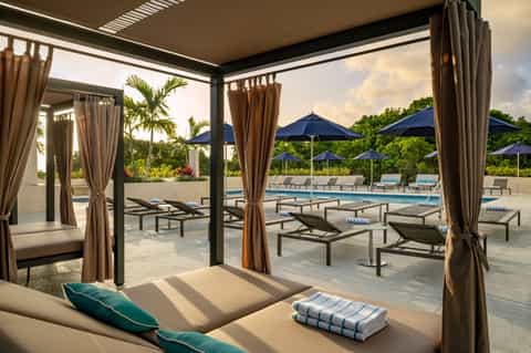 Covered poolside cabana with lounge beds, sheer curtains, and view of resort pool with umbrellas beyond