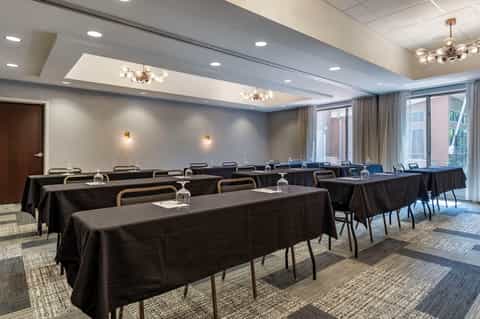 Empty meeting room with rows of black-covered tables, chairs, exposed beam ceiling, and large windows