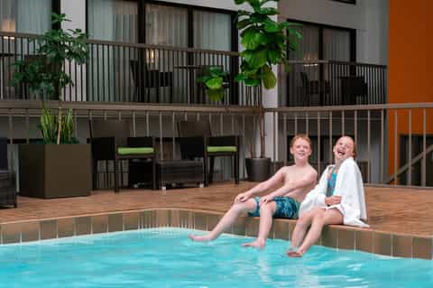 Two children sitting poolside on wooden deck with modern hotel balconies and green accent walls visible