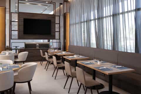 Contemporary restaurant interior with wood wall panel, flat-screen TV, white chairs, dining tables, and sheer curtains