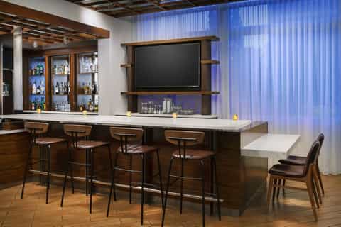 Modern hotel bar with wooden cabinetry, flat-screen TV, barstool seating, and blue curtains