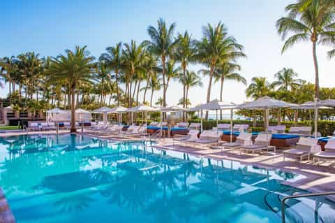 Beachfront pool with turquoise water, white umbrellas, palm trees, and pastel loungers lining the deck