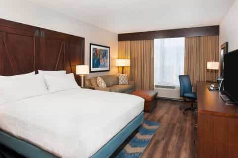 Hotel bedroom with queen bed, dark wood headboard, seating area, and city window views