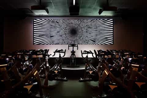Indoor spin studio with rows of exercise bikes facing large illuminated geometric wall display screen