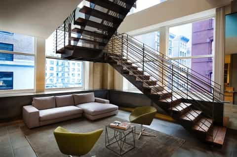Industrial loft-style lobby with floating staircase, metal railings, large windows overlooking city skyline, and modern seating