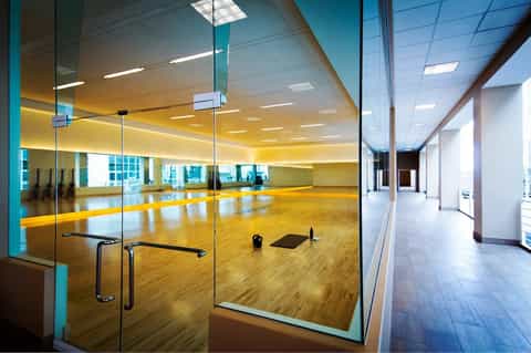 Modern fitness studio with mirrored walls, wooden floor, glass doors, yellow lighting, and city views