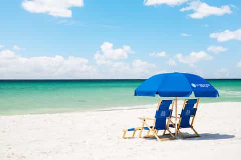 Beach setup with blue umbrella and two beach chairs on white sand overlooking turquoise ocean