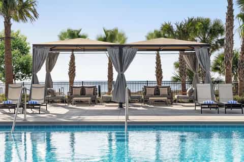 Resort pool with dual gray pergolas, lounge seating, palm trees, and ocean waterfront railing