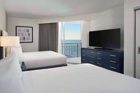 Oceanview hotel bedroom with two queen beds, balcony, TV, and coastal horizon views