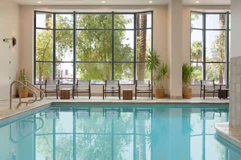 Indoor pool with large windows overlooking palm trees, wooden seating area, and natural light