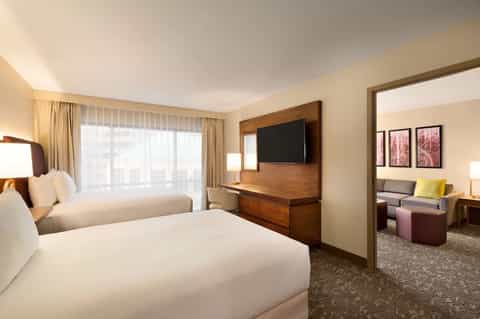 Spacious hotel bedroom with two queen beds, wooden TV wall, sitting area, and floor-to-ceiling windows
