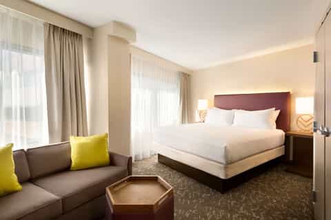 Hotel bedroom with king bed, burgundy headboard, sofa with yellow pillow, and windows with sheer curtains
