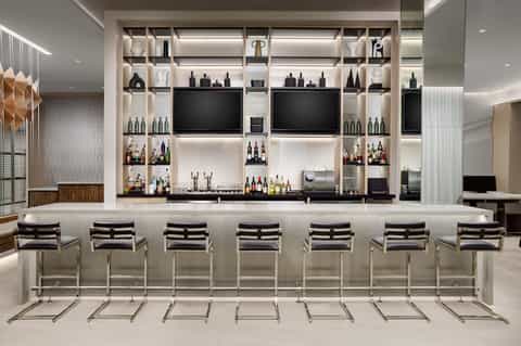 Contemporary hotel bar with white shelving, bottles of wine and spirits, flat-screen TVs, and black bar stools