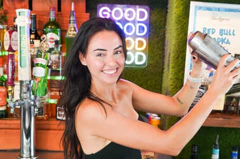 Woman mixing cocktail at tropical outdoor bar with neon good mood sign and greenery backdrop