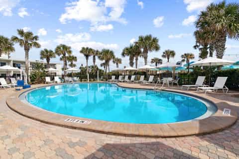 Curved community pool surrounded by palm trees, white umbrellas, and lounge chairs on brick deck.