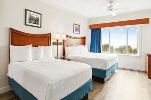 Hotel bedroom with two queen beds, wooden headboards, ocean view windows, and blue curtains