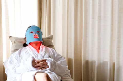 Person wearing white robe and blue mask relaxing in chair with light curtains in spa or resort room