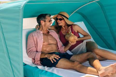 Couple relaxing in turquoise beach cabana with shade, man and woman enjoying drinks poolside