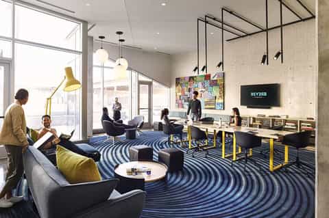Contemporary hotel lounge with blue patterned carpet, seating areas, work tables, and bright windows